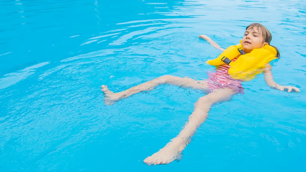 A small boy floating on his back in a pool, arms relaxed at his sides, under a bright sky.