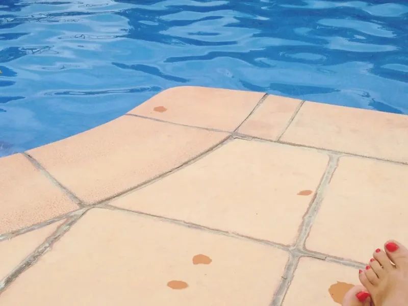 Young woman relaxing by modern pool border with legs in water during sunny vacation day