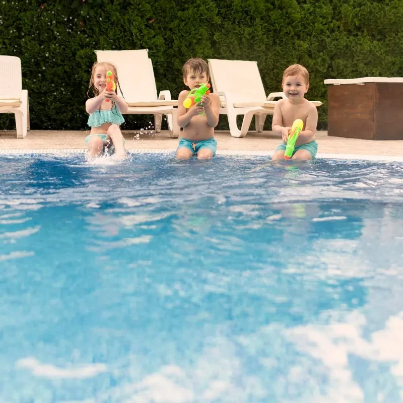 Three children laughing and playing with water guns in a sparkling blue backyard swimming pool on a sunny day