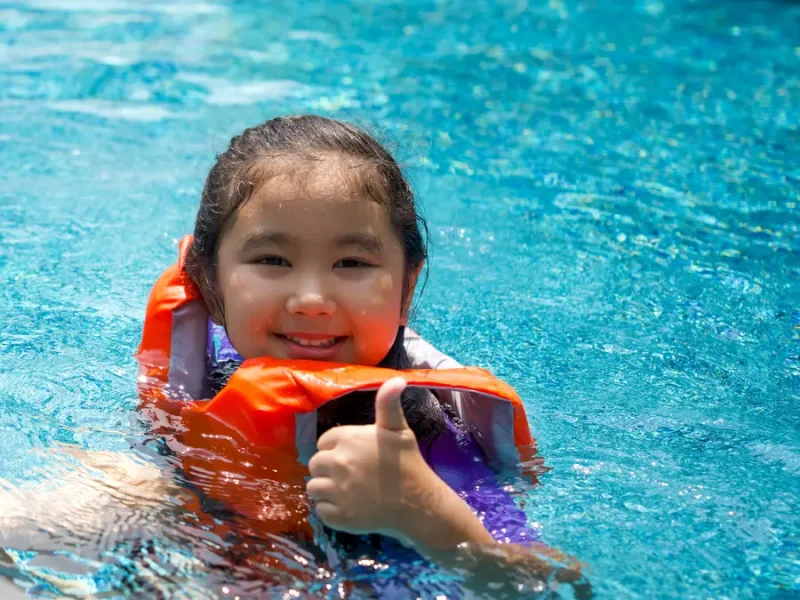 Kid in orange float jacket giving a thumbs up while enjoying the pool